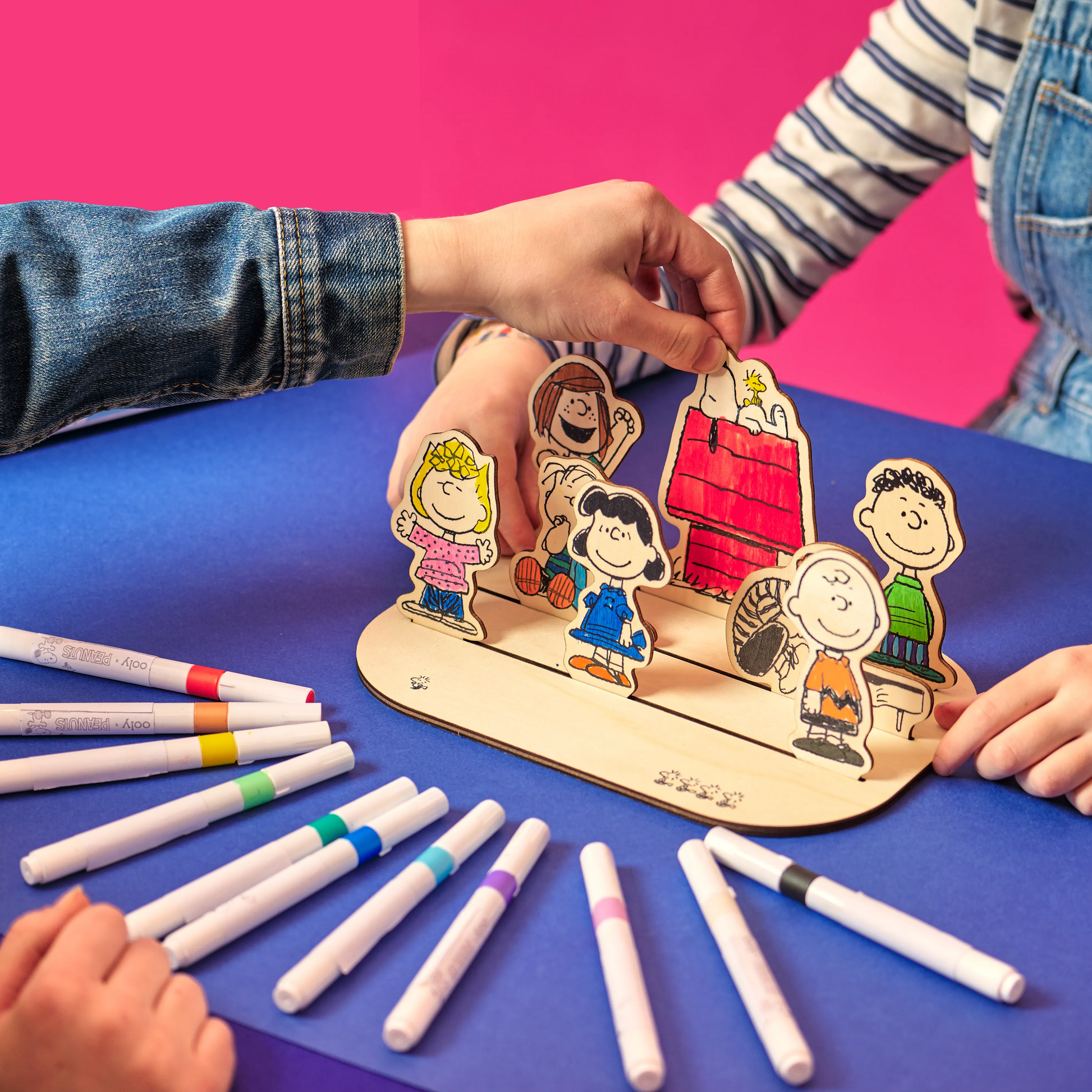 Two people setting up the OOLY x Peanuts Color-In Wooden Scene with paint markers