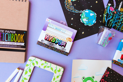 OOLY Vivid Pop! Metallic Paint Markers on a purple table with art and supplies