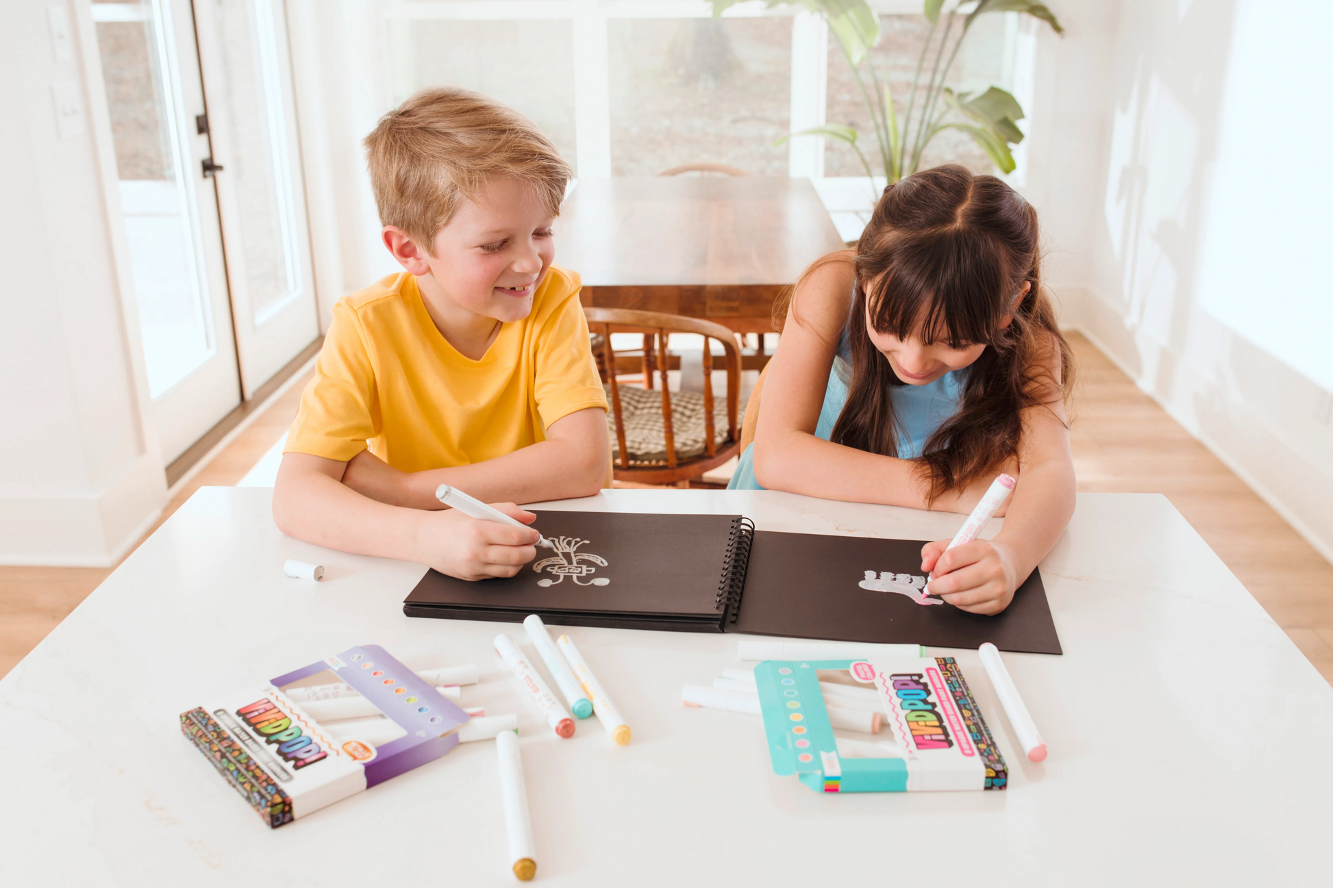 Two kids coloring on black paper pad with OOLY Vivid Pop! Paint Markers wide shot